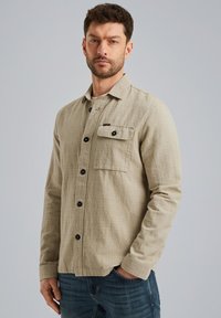 Beige gevlochten shirt met een button-down kraag, lange mouwen, zwarte knopen en een enkele borstzak, gedragen over een paar blauwe spijkerbroeken.
