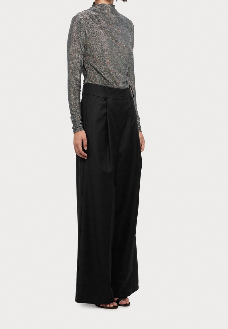Langärmliges Oberteil aus metallisch grauem Stoff mit einem drapierten Design; kombiniert mit lockeren schwarzen Hosen, die einen weiten Bein und hohe Taille haben.