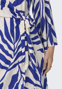 Wikkeljurk van crème stof met gedurfde blauwe bladerenpatronen, met een geknoopt detail in de taille en brede mouwen.