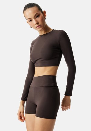 Kvinde iført matchende mørkebrun langærmet crop top og højtaljede tætsiddende shorts, stående foran en ensfarvet lys baggrund.