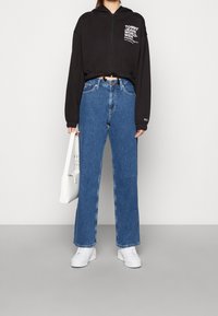 Sudadera negra corta con texto blanco, jeans azules de talle alto, zapatillas blancas y un bolso blanco pequeño. Fondo liso.