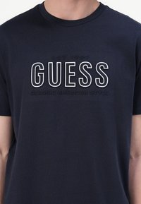 Navy blue cotton t-shirt featuring raised white text "GUESS EST 1981 CLASSIC AMERICAN STYLE" centered on the chest. Short sleeves, crew neck.