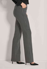 Vrouw in zwart-wit gestreepte wijdlopende broek met zwarte elegante puntige hakken, hand in haar zak, staand tegen een lichte muur.