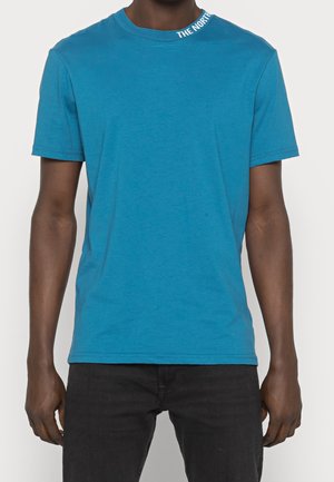 Mężczyzna noszący jednokolorowy turkusowy T-shirt z krótkim rękawem z napisem "THE NORTH" po lewej stronie kołnierza oraz czarne spodnie.