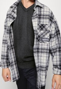 Chemise en flanelle à carreaux noir et blanc avec une seule poche poitrine, portée sur un pull gris à col en V. Tissu doux et texturé.