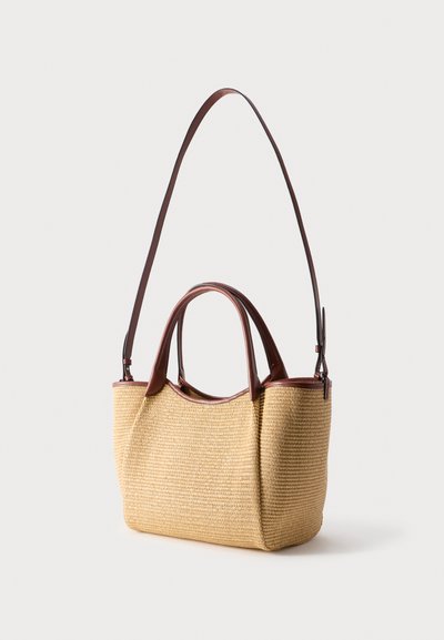 Πλεκτή μπεζ τσάντα με καφέ δερμάτινες λαβές και ρυθμιζόμενο λουράκι ώμου σε ουδέτερο φόντο.