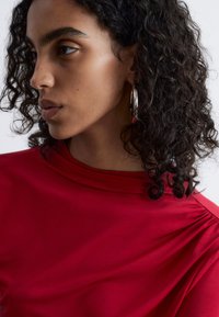 Rotes Top mit hohem Kragen und drapierten Details, aus glattem Stoff gefertigt. Hat lockige Haare und große silberne Creolen.