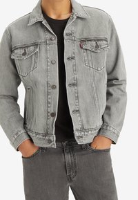 Graue Jeansjacke mit zwei Brusttaschen, Metallknöpfen und einem Kragen. Getragen über einem schwarzen T-Shirt, kombiniert mit grauen Jeans.