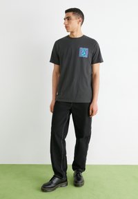 Una camiseta de algodón gris oscuro presenta un gráfico geométrico colorido en el pecho; combinada con pantalones negros y zapatos negros gruesos.