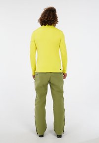 Man met krullend haar, gekleed in een felgeel shirt met lange mouwen en olijfgroene broek, staand met zijn rug naar de camera op een witte achtergrond.