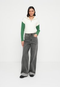 Biela a zelená sveter so zipsom v kombinácii s vysokými širokými šedými džínsami. Džínsové nohavice majú jemne ošúchaný vzhľad a vyhrnuté manžety.