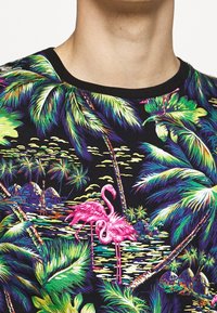 Buntes tropisches Muster mit zwei pinken Flamingos, grünen Palmen und Hütten auf schwarzem Hintergrund, dargestellt auf dem Oberkörper einer Person.