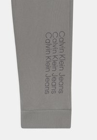 Szare spodnie sportowe z prążkowanymi mankietami, z powtarzającym się napisem „Calvin Klein Jeans” w ciemniejszym odcieniu na boku. Gładka faktura.
