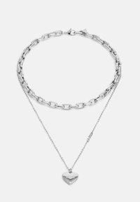 Collier en argent avec un design en couches. Il présente un style de maillons chunky et une chaîne délicate avec un pendentif en forme de cœur portant le mot "GUESS."