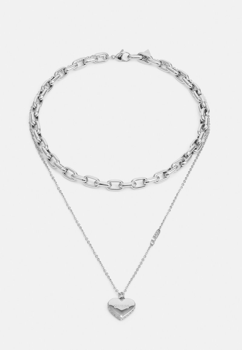 Collier en argent avec un design en couches. Il présente un style de maillons chunky et une chaîne délicate avec un pendentif en forme de cœur portant le mot "GUESS."