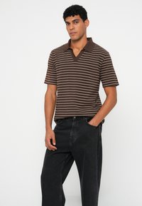 Man wearing brown and white striped short-sleeve polo shirt and loose black jeans, standing with one hand in pocket against white background.