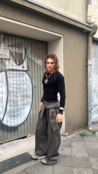 Juoda ilgomis rankovėmis marškiniai su žvaigždės pakabučiu, pilkos dryžuotos plačios kelnės, raštuota medžiaga apvyniota aplink liemenį ir pilkos avalynės.