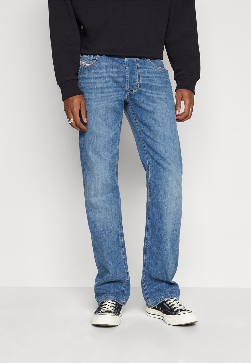 Jean en denim bleu clair, coupe droite, couture visible et légère décoloration. Portés avec des baskets noires à semelles blanches.