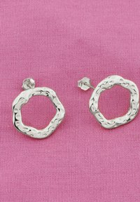 Silverfärgade rundörhängen med en oregelbunden yta, med ett utskuret mönster. Positionerade mot en rosa tygbakgrund.