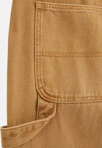 Beige Stoff mit strukturiertem Oberflächen, ausgestattet mit einer aufgesetzten Tasche und einem umgeschlagenen Saum. Verstärkte Nähte entlang der Kanten und ein robustes Design.