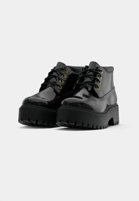 Scarpe in vernice nera con punta tonda, allacciatura frontale e suole in gomma nera spessa con un motivo di battistrada robusto.