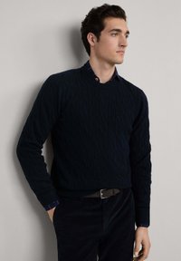 Maglione a trecce blu navale con maniche lunghe, scollo rotondo e polsini a costine, indossato sopra una camicia a quadri blu, abbinato a pantaloni scuri.