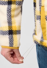Giacca in fleece con un motivo plaid giallo, blu navy e grigio. La mano tira un cordino dall'orlo. I polsini presentano una fascia gialla a contrasto.
