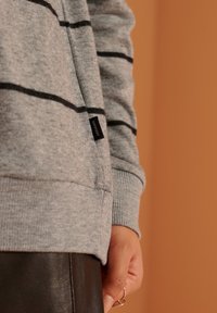 Felpa grigia con strisce nere, polsini a coste e un'etichetta che recita "Superdry". Il tessuto sembra morbido e ha una consistenza liscia.