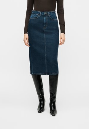 Femme portant une jupe crayon en denim bleu foncé avec des poches avant et des bottes noires montantes, debout devant un fond blanc uni.
