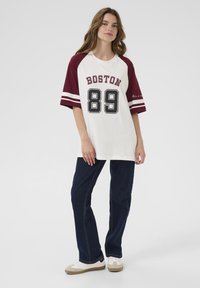 Bílé a bordó oversized tričko s potiskem "BOSTON 89", kontrastními rukávy a pruhy. V kombinaci s tmavě modrými rovnými džíny a světlejšími botami.