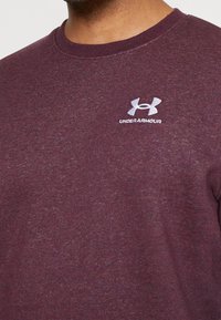 Homme portant un sweat-shirt à col rond Under Armour couleur bordeaux avec un logo blanc brodé sur la poitrine.