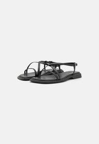 Zwarte platte sandalen met meerdere overlappende banden, een gladde textuur en een gesp-sluiting. De zool is iets verhoogd en rechthoekig.