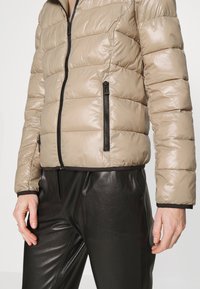Beżowa kurtka puchowa z poziomymi stębnowaniami, czarnym zamkiem błyskawicznym i prążkowanymi mankietami, zestawiona z błyszczącymi, czarnymi skórzanymi spodniami.