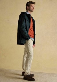 Jachetă impermeabilă verde închis cu glugă, pulover portocaliu, cămașă cu carouri, pantaloni din corduroy crem și pantofi din piele maro cu șireturi, stând pe o suprafață texturată.