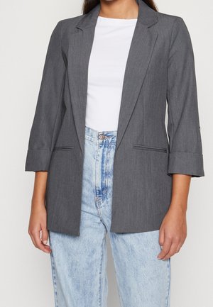 Femme portant un blazer gris à manches trois-quarts sur une chemise blanche et un jean taille haute bleu clair.