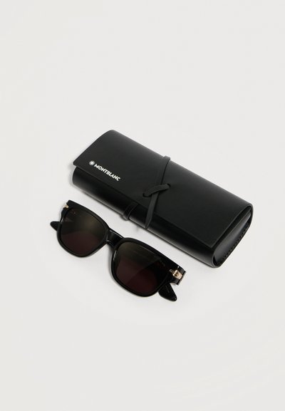 Μαύρα ορθογώνια γυαλιά ηλίου με σκουρόχρωμους φακούς δίπλα σε μαύρη δερμάτινη θήκη Montblanc δεμένη με λουράκι σε απλό λευκό φόντο.