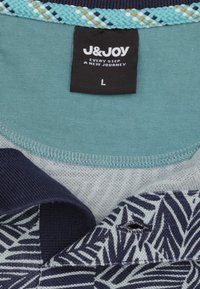 Kortebloes met teal en marineblauwe kleuren, voorzien van een bladerenpatroon in marineblauw en een textuurrijk gevlochten kraag. Het etiket toont het merk en de maat.