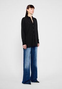 Camicia nera a maniche lunghe con bottoni in tessuto testurizzato, abbinata a jeans in denim blu a gamba larga con un effetto di colore sfumato e scarpe nere a punta.