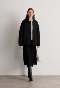 Zwarte oversized bomberjack met een gladde textuur, gedragen over een witte top. Gecombineerd met een zwarte geplooide rok en zwarte kniehoge laarzen.