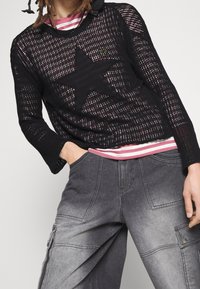 Schwarzer Strickpullover mit einem großen Sternmotiv, über einem rosa-weiß gestreiften Hemd getragen, kombiniert mit grauen Cargo-Hosen mit Taschen.