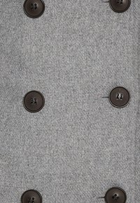 Tissu gris à finition texturée, avec six boutons bruns et un design croisé. Les boutons ont deux trous chacun.