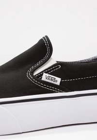 Scarpe Vans nere slip-on con cuciture bianche, dettagli elastici sui lati e suola in gomma bianca su uno sfondo bianco semplice.