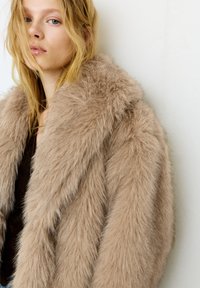 Femme blonde aux cheveux lâchés portant un grand manteau en fourrure beige duveteux, debout contre un mur blanc texturé.