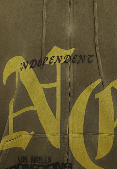 Olívovo zelená mikina na zips vyrobená z bavlny, s veľkým žltým grafickým textom a akcentmi. Obsahuje klokaní vrecko a šnúrku na kapucni.