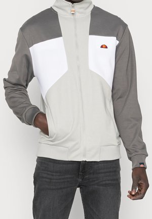 Homme portant une veste zippée avec des manches grises, un devant gris clair, un panneau poitrine blanc et un petit logo rouge et orange sur la poitrine et le poignet.