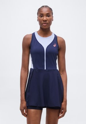 Femme aux cheveux tressés portant une robe de sport sans manches bleu marine et bleu clair avec un logo ASICS sur la poitrine, debout devant un fond blanc uni.