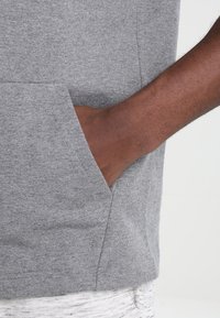 Mano oscura apoyada dentro del bolsillo de un sudadera gris, con parte de un pantalón de color claro visible abajo.