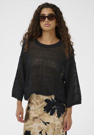 Frau mit lockigem Haar, die eine Schildpatt-Sonnenbrille, ein lockeres schwarzes Strickoberteil und einen beigen Rock mit Blumendruck trägt.