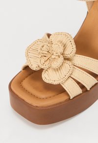 Beige plattform sandaler med et vevd design, prydet med en teksturert blomsterdetalj og flere stropper, satt på en brun base.