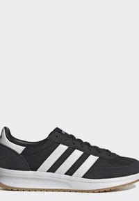Zapatilla Adidas en blanco y negro con tres rayas blancas, cordones negros, suela blanca acolchada y suela exterior de goma sobre un fondo gris claro.